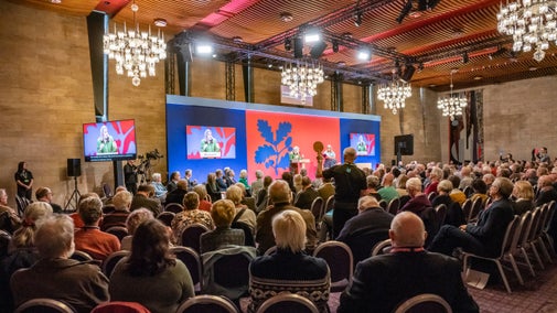 A seated audience watch a lady speaking at a podium in front of a brightly coloured backdrop with chandeliers overhead